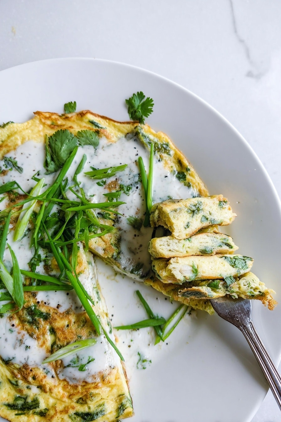 A white plate with a layered omelet that has green herbs visible inside, folded into sections showing a bumpy yellow texture with green flecks. The top layer is covered with a smooth white sauce and garnished with fresh green cilantro leaves and long thin green onion pieces placed on top. A piece of the omelet is being lifted on a silver fork, showing the soft and slightly browned interior. The background is a white marbled surface. photo taken with an iphone --ar 2:3 --v 7