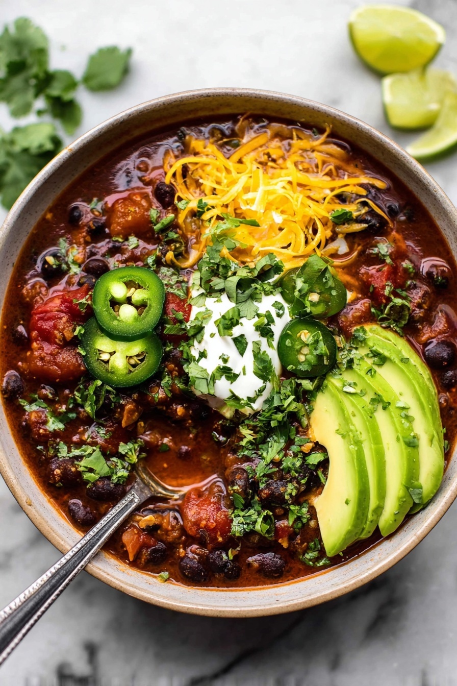 A deep bowl filled with rich, dark chili containing visible black beans and small chunks of red tomatoes in a thick red sauce. On top, there are three slices of green jalapeño and several slices of light green avocado fanned out on the right side. A generous sprinkle of shredded yellow cheddar cheese lies on the upper left, with a dollop of white sour cream garnished with finely chopped green onion in the center. Fresh green cilantro leaves and chopped herbs are scattered across the chili, adding texture and color. A spoon dipped into the chili shows the thick mixture inside. The bowl sits on a white marbled surface with three slices of lime in the upper right. Photo taken with an iphone --ar 2:3 --v 7
