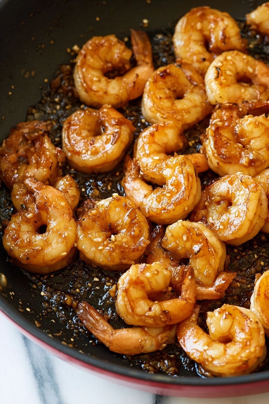 A close-up view of cooked shrimp in a red skillet, showing about two layers of shrimp piled together, each shrimp curled into a crescent shape with a shiny, glazed surface that is light orange and brown. The skillet bottom is black with some bits and a slight oily texture, contrasting with the shrimp. The skillet sits on a white marbled surface. Photo taken with an iphone --ar 2:3 --v 7