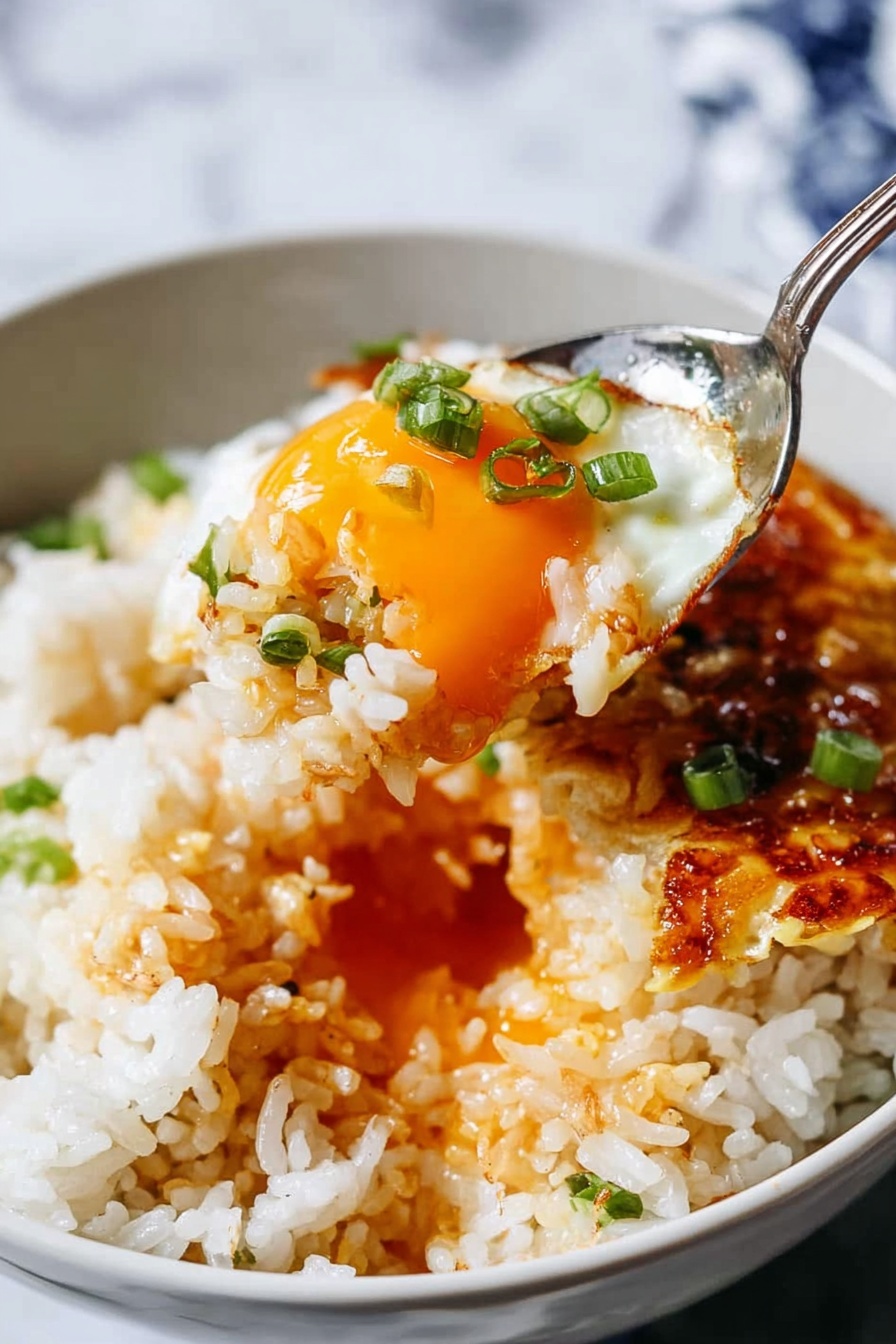 A close-up image of a white bowl filled with cooked white rice topped with a fried egg that has a golden crispy edge and a runny orange yolk in the center. The rice shows some bits of light brown crispy edges mixed in. Small pieces of green onions are sprinkled over the rice. A spoon is lifting a portion of the rice mixed with the egg yolk, showing a glistening texture with bits of green onion on top. The background has a white marbled surface. photo taken with an iphone --ar 2:3 --v 7